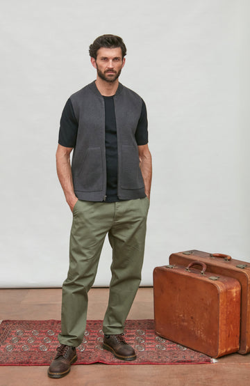 A man stands in a studio wearing a dark gray sleeveless zip-front vest over a black short-sleeve shirt, olive green pants, and brown shoes. A vintage brown suitcase and a red patterned rug are on the floor against a light gray backdrop.  Knitted in Cotton by Paul James Knitwear.