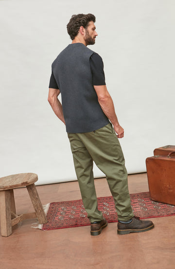 A man stands with his back to the camera wearing a dark charcoal gray short-sleeve knit top and olive green pants, standing in a styled studio setting with a wooden stool, rug, and leather suitcase.  Knitted in Cotton by Paul James Knitwear.