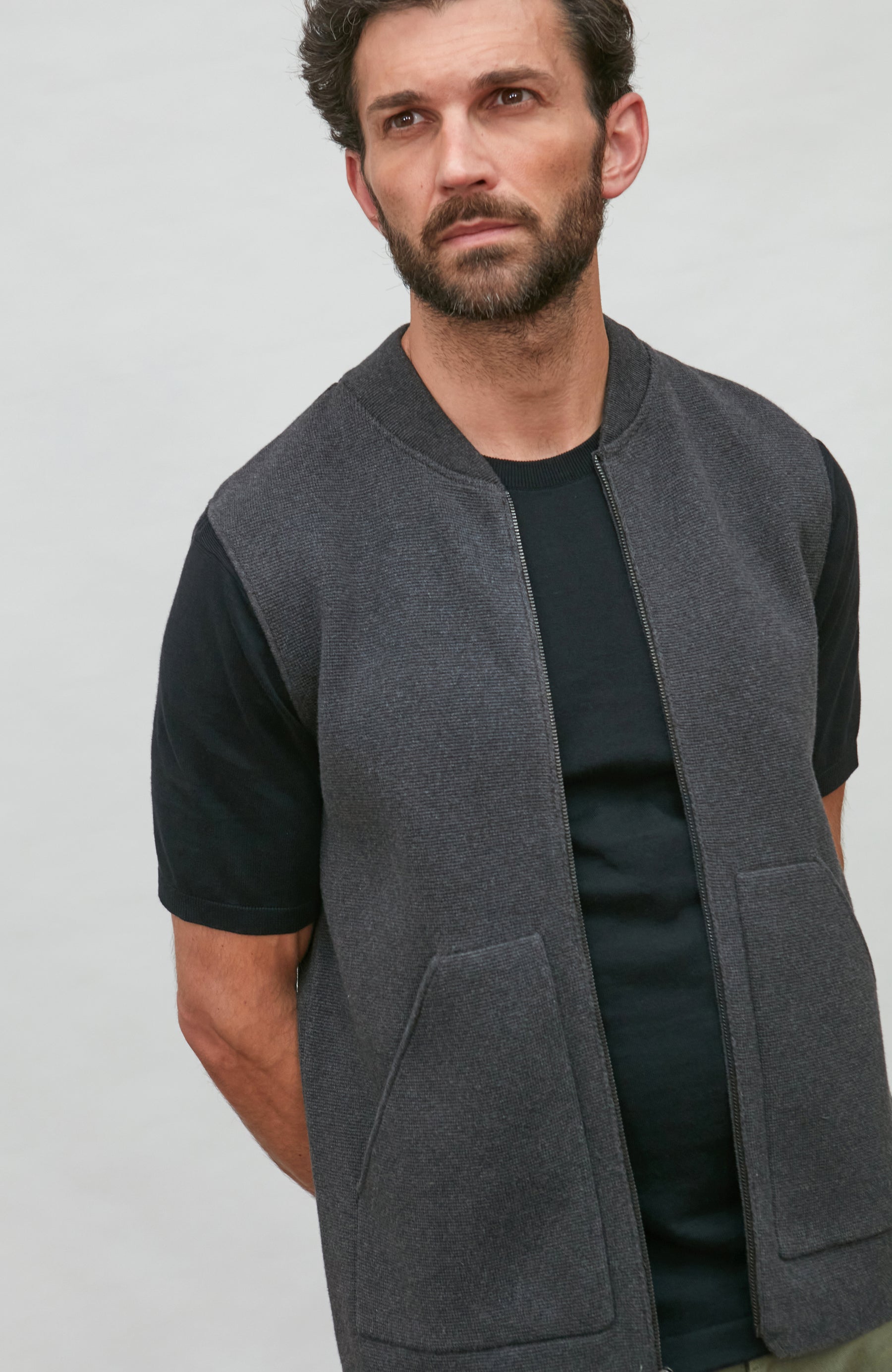 A man wearing a dark gray sleeveless zip-front vest over a black t-shirt, photographed against a plain light gray studio background. Knitted in Cotton by Paul James Knitwear. 