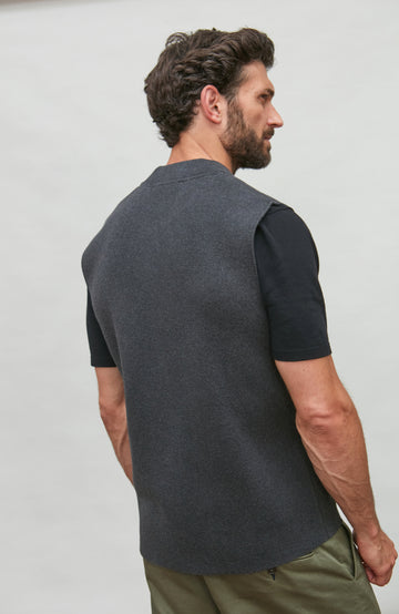 A man shown from the back wearing a dark gray knit sleeveless vest over a black short-sleeve shirt and olive green pants, against a plain light gray background.  Knitted in Cotton by Paul James Knitwear.
