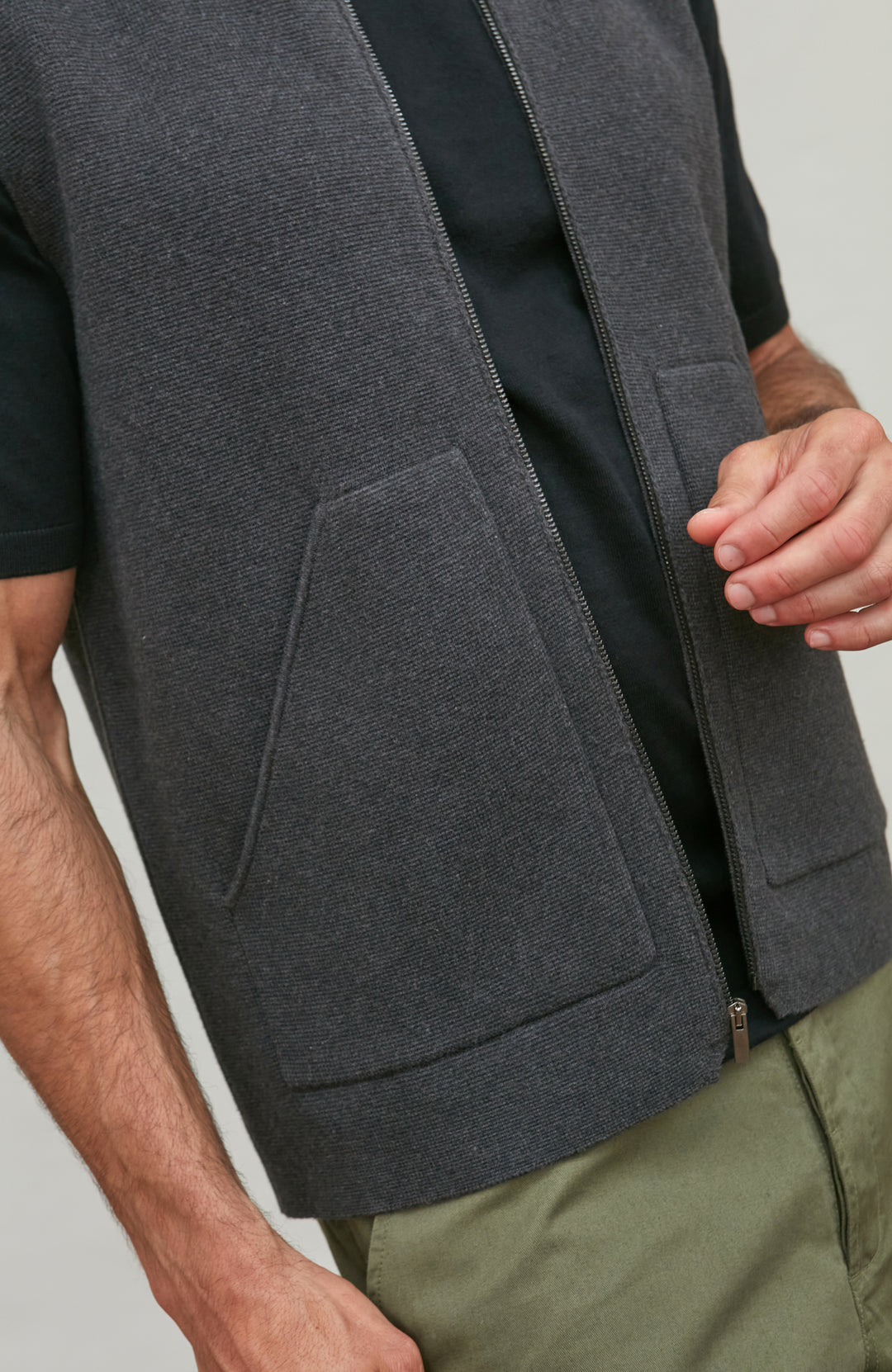A close-up of a person wearing a dark gray zip-front vest over a black short-sleeve shirt and olive green pants, against a light gray studio background.  Knitted in Cotton by Paul James Knitwear.