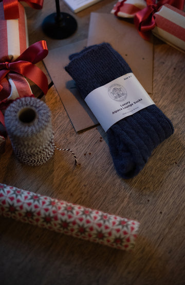 Pair of navy alpaca socks with a label on a wooden surface surrounded by gift ribbons and wrapping paper.