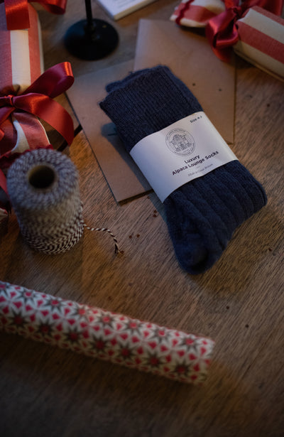 Pair of navy alpaca socks with a label on a wooden surface surrounded by gift ribbons and wrapping paper.