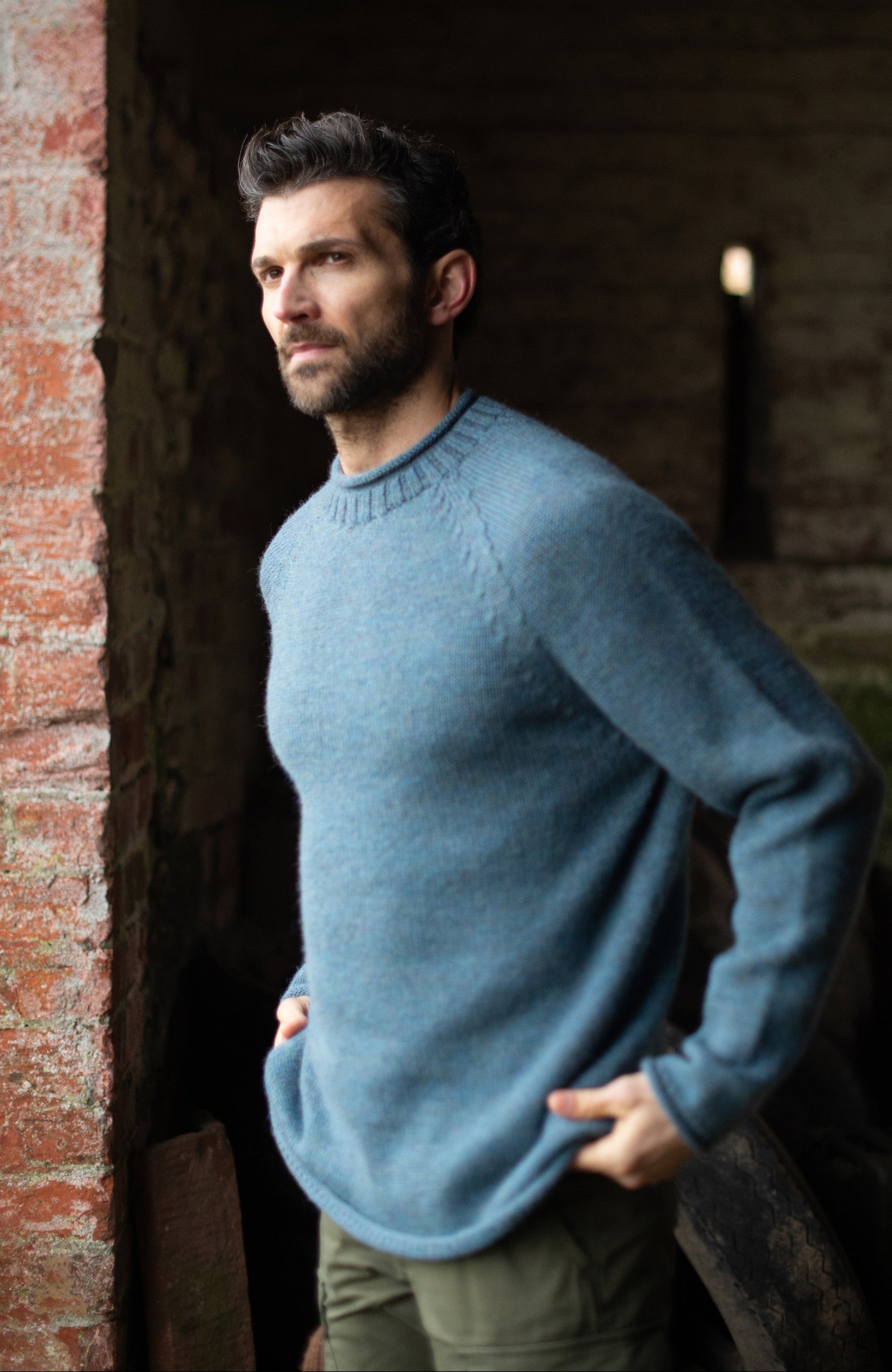 A man wearing a made in england dusty blue knit british wool sweater by paul james knitwear and olive green pants, standing in a rustic brick-walled corridor.