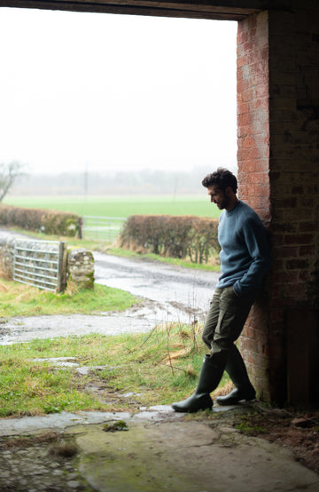 A man wearing a made in england dusty blue knit british wool sweater by paul james knitwear and olive green pants, standing in a rustic brick-walled corridor.