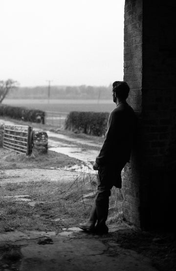 A black and white photograph of a man in silhouette standing in the shadow of a brick wall, looking out onto a rural field and fence by pal james knitwear