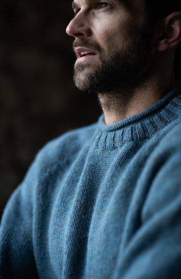 A man wearing a made in england dusty blue knit british wool sweater by paul james knitwear and olive green pants, standing in a rustic brick-walled corridor.