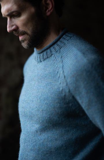 A close-up side profile of a man wearing a made in england British wool fisherman textured knit crewneck sweater by paul james knitwear in a muted dusty blue color, set against a dark, moody background.