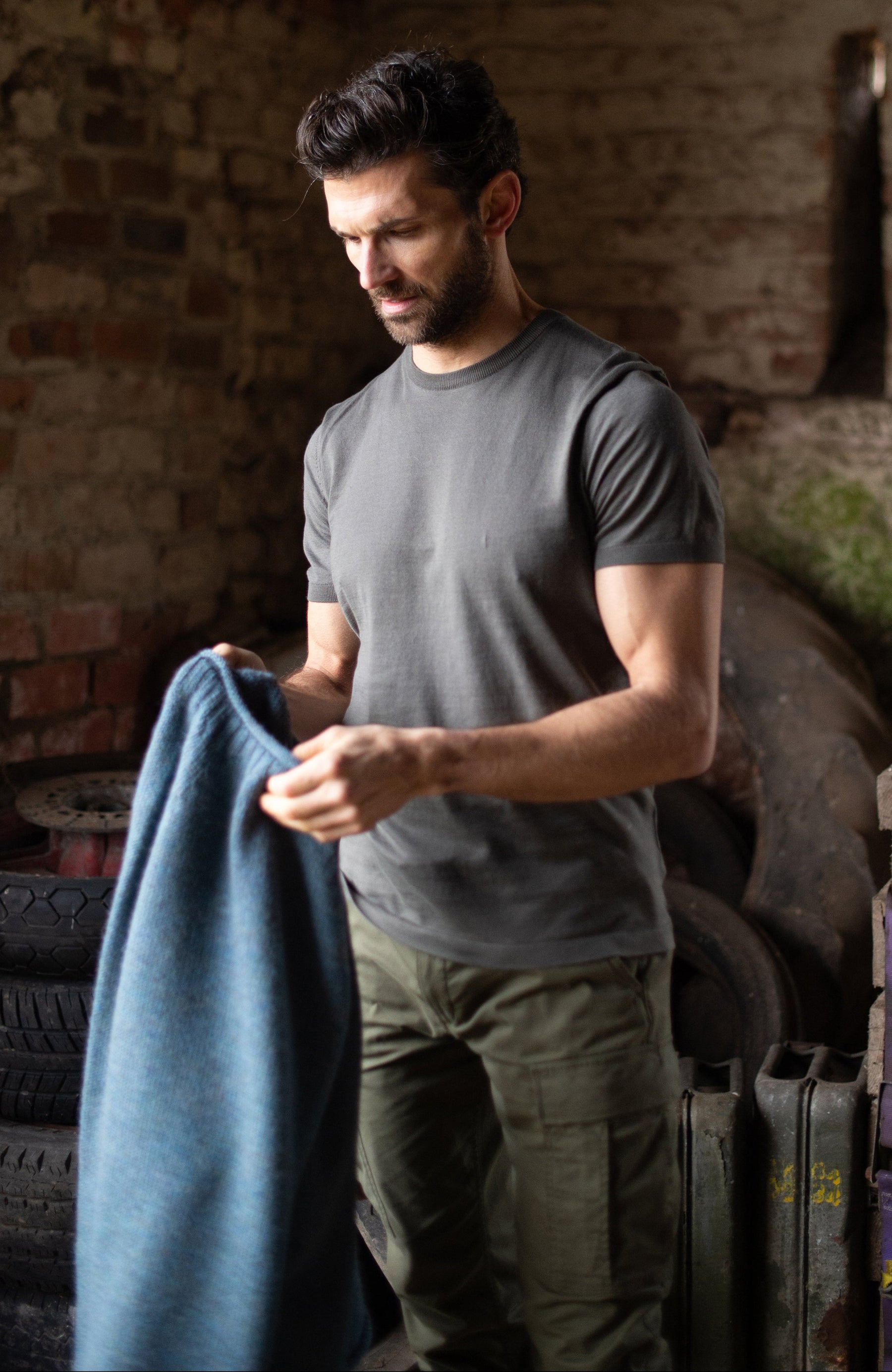 A man in a gray mercerised cotton t-shirt by Paul James Knitwear and green pants stands in a rustic brick room, holding a folded blue-gray blanket or towel.