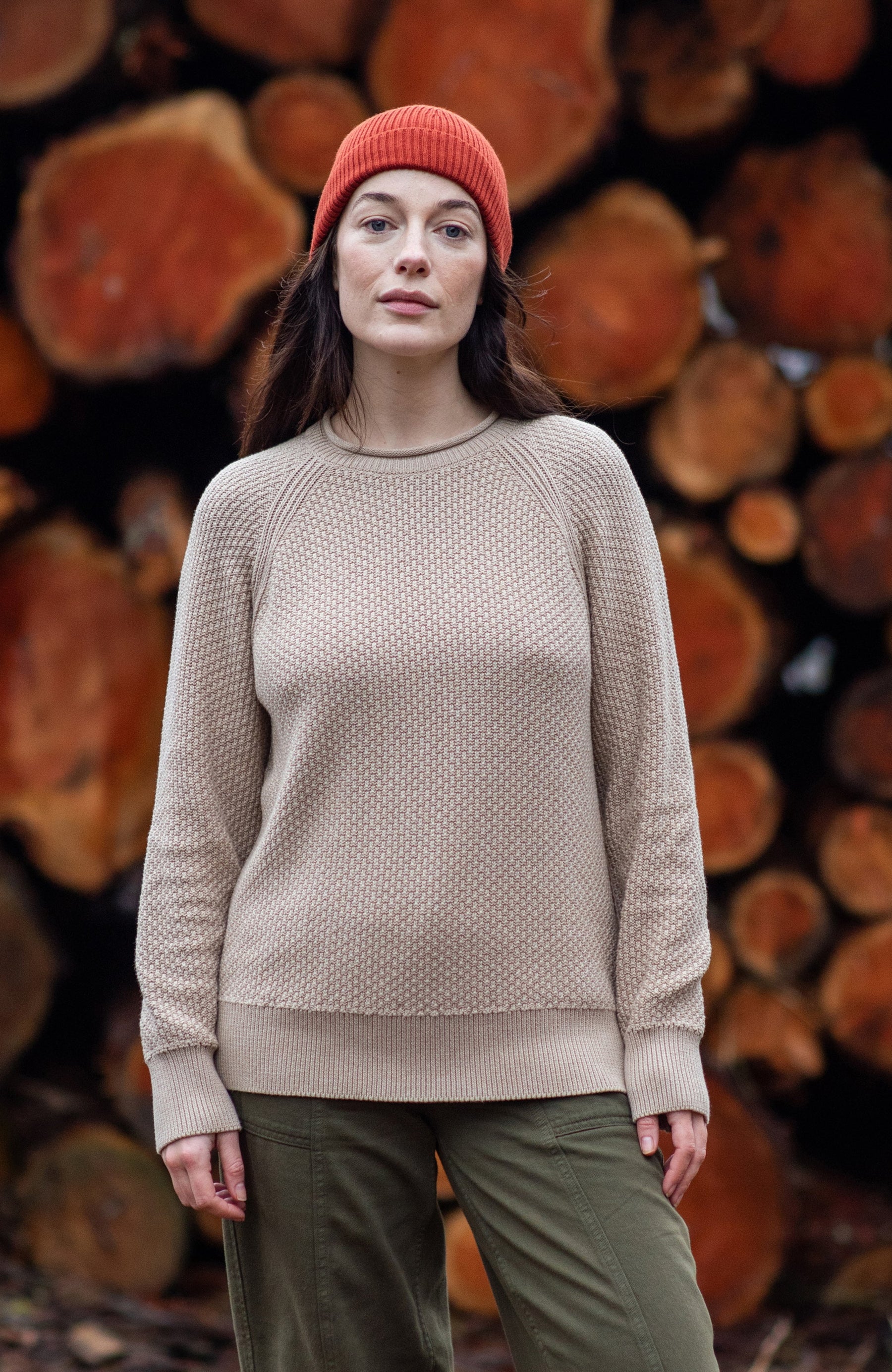 Fisherman Jumpers - A woman stands outdoors in front of a large stack of cut logs, wearing a beige knit sweater by paul james knitwear, olive green pants, and a rust-orange beanie. 
