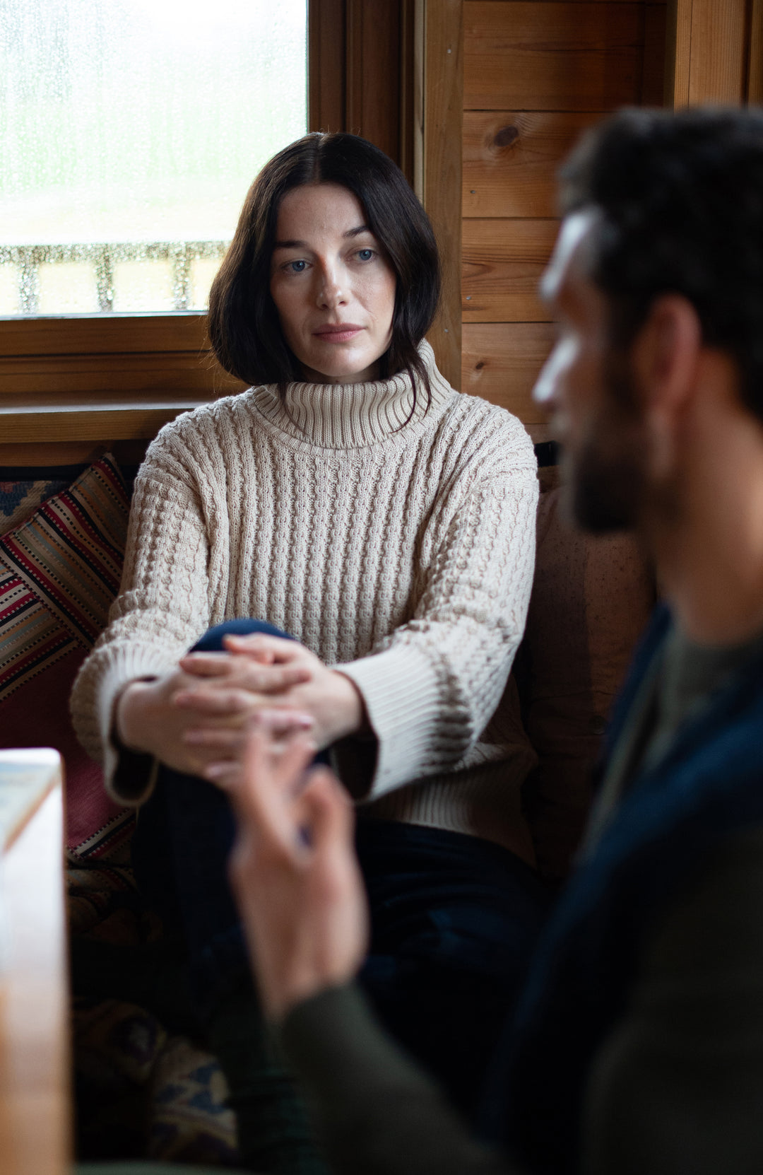 A woman wearing a cream cable-knit turtleneck cotton sweater by Paul James Knitwear sits in a rustic wooden room, looking thoughtfully toward a man in the foreground who is out of focus. 