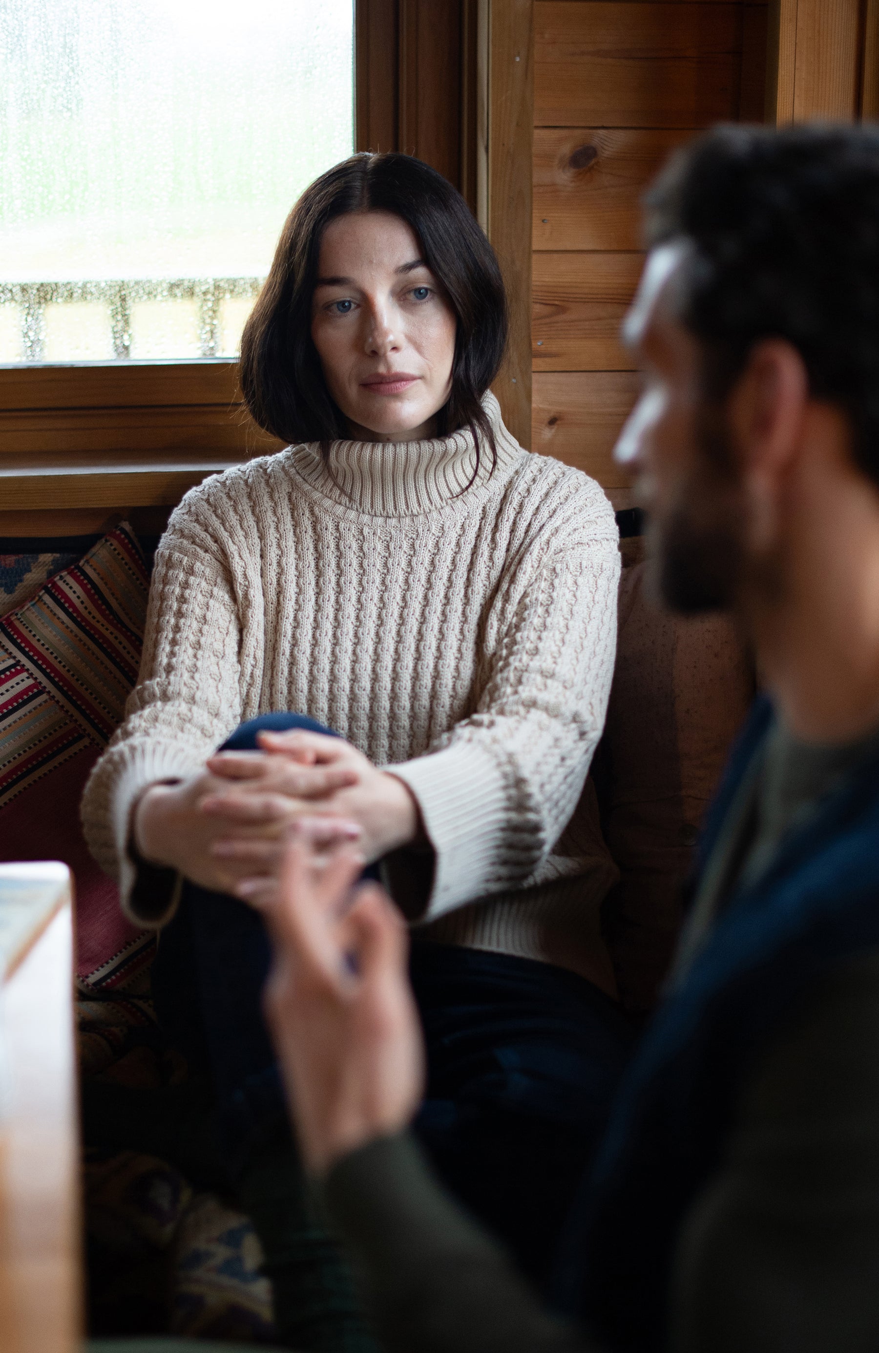 A woman wearing a cream cable-knit turtleneck cotton sweater by Paul James Knitwear sits in a rustic wooden room, looking thoughtfully toward a man in the foreground who is out of focus. 