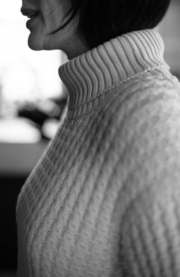 Close-up side profile of a person wearing a chunky ribbed turtleneck sweater in a black-and-white made by Paul James Knitwear photograph.