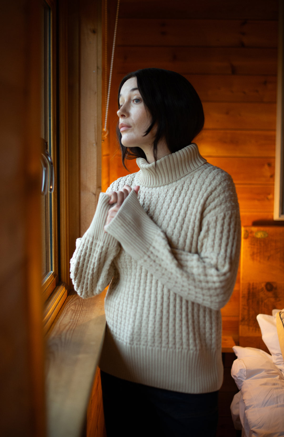 A woman with short dark hair wearing a cream-colored Cotton Paul James Knitwear ribbed turtleneck sweater stands by a window in a warm wood-paneled room.