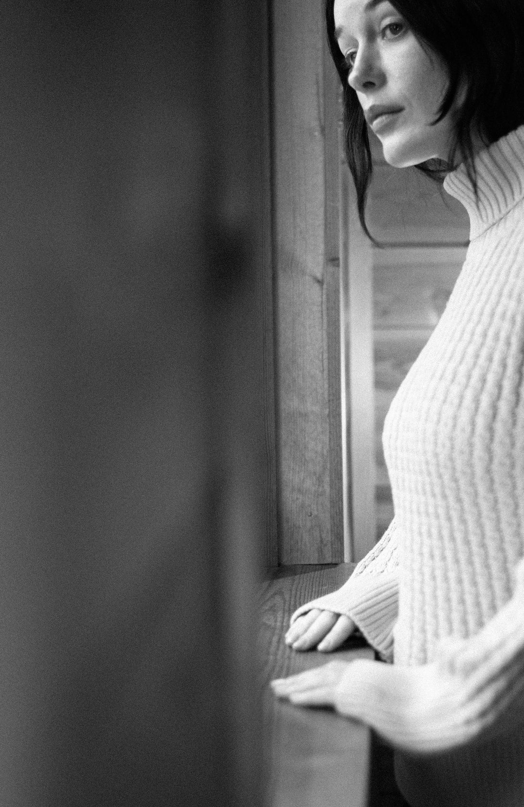 A black and white portrait of a woman in a ribbed turtleneck sweater made by Paul James Knitwear leaning against a window.