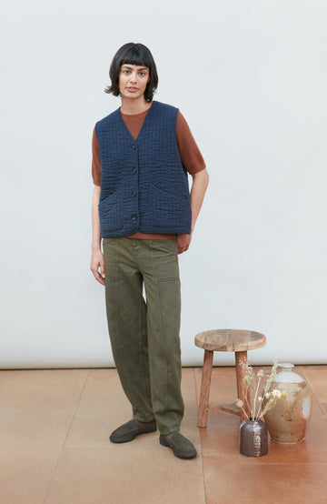 A women wearing a navy blue quilted button-up vest by Paul James Knitwear over a rust-brown short-sleeve top and olive green pants, photographed against a light gray studio background.