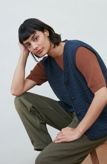A women wearing a navy blue quilted button-up vest by Paul James Knitwear over a rust-brown short-sleeve top and olive green pants, photographed against a light gray studio background.
