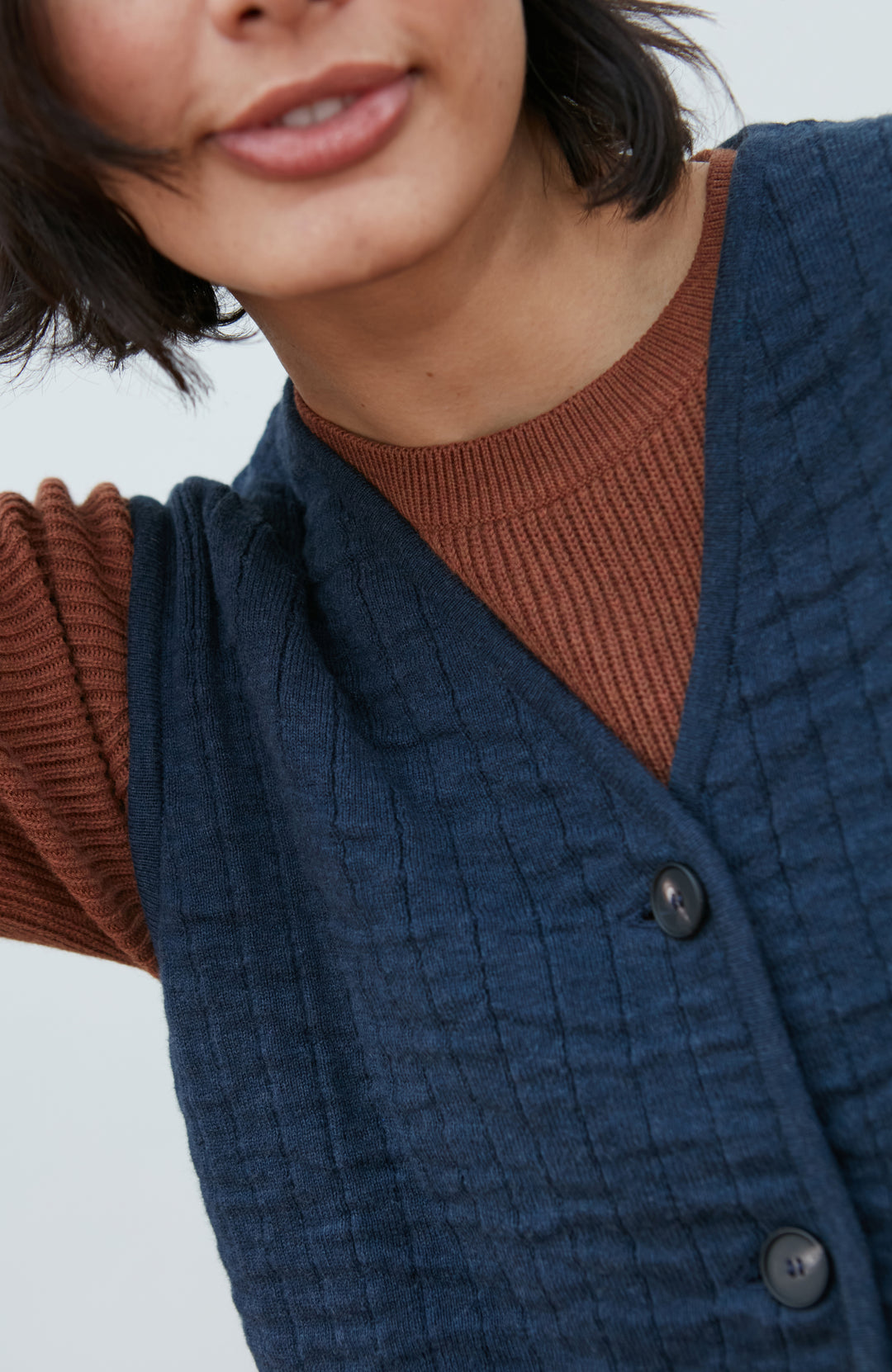 A women wearing a navy blue quilted button-up gilet by Paul James Knitwear over a rust-brown short-sleeve top and olive green pants, photographed against a light gray studio background.