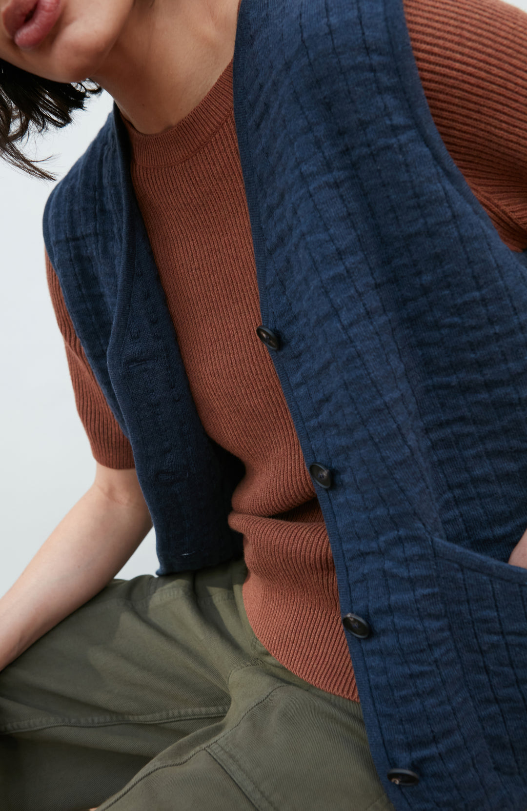 A women wearing a navy blue quilted button-up gilet by Paul James Knitwear over a rust-brown short-sleeve top and olive green pants, photographed against a light gray studio background.