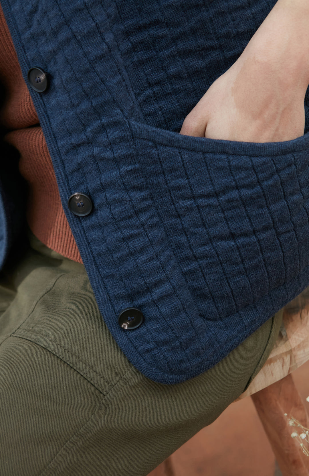 A women wearing a navy blue quilted button-up gilet by Paul James Knitwear over a rust-brown short-sleeve top and olive green pants, photographed against a light gray studio background.