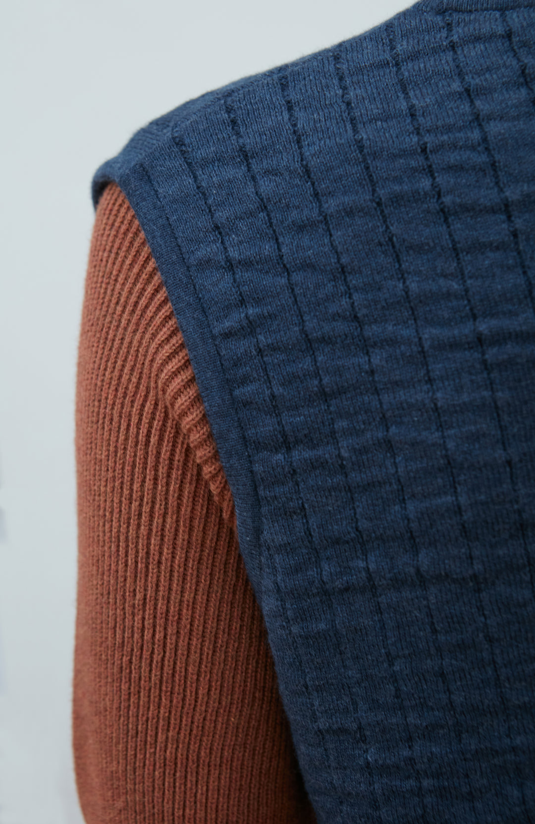 A women wearing a navy blue quilted button-up gilet by Paul James Knitwear over a rust-brown short-sleeve top and olive green pants, photographed against a light gray studio background.