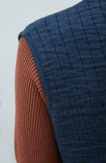 A women wearing a navy blue quilted button-up gilet by Paul James Knitwear over a rust-brown short-sleeve top and olive green pants, photographed against a light gray studio background.