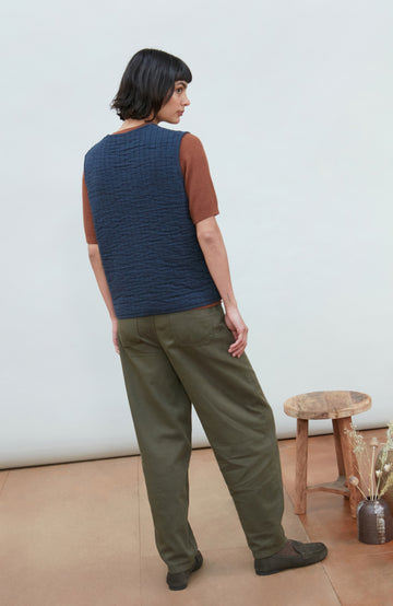 A women wearing a navy blue quilted button-up gilet by Paul James Knitwear over a rust-brown short-sleeve top and olive green pants, photographed against a light gray studio background.