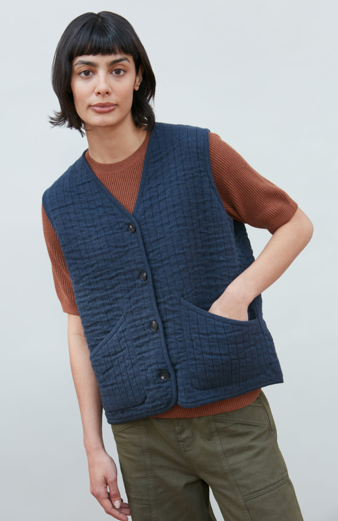 A women wearing a navy blue quilted button-up vest by Paul James Knitwear over a rust-brown short-sleeve top and olive green pants, photographed against a light gray studio background.