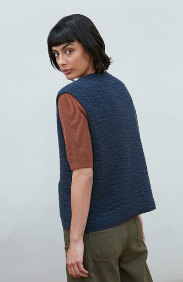 A women wearing a navy blue quilted button-up gilet by Paul James Knitwear over a rust-brown short-sleeve top and olive green pants, photographed against a light gray studio background.