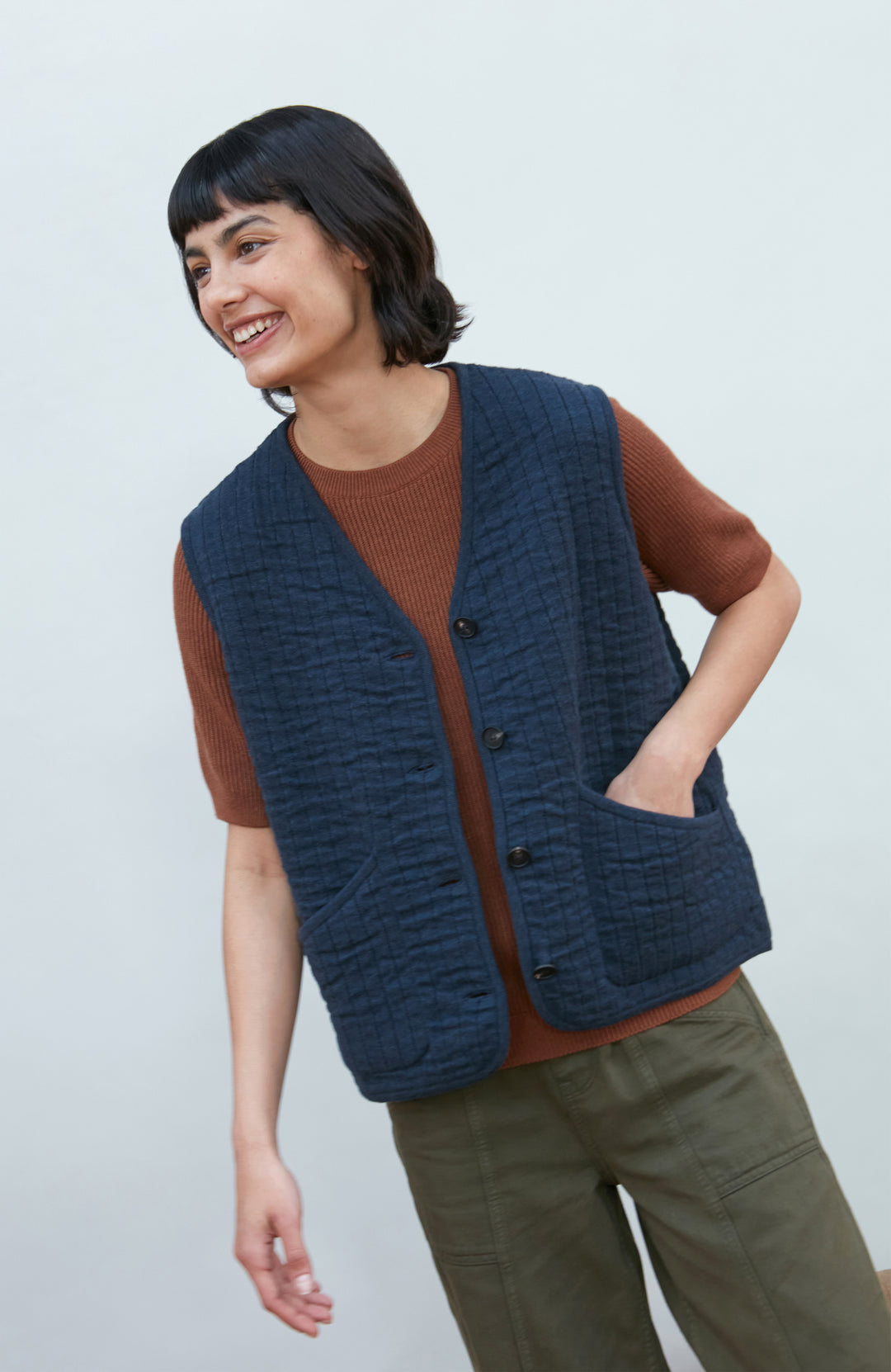 A women wearing a navy blue quilted button-up vest by Paul James Knitwear over a rust-brown short-sleeve top and olive green pants, photographed against a light gray studio background.