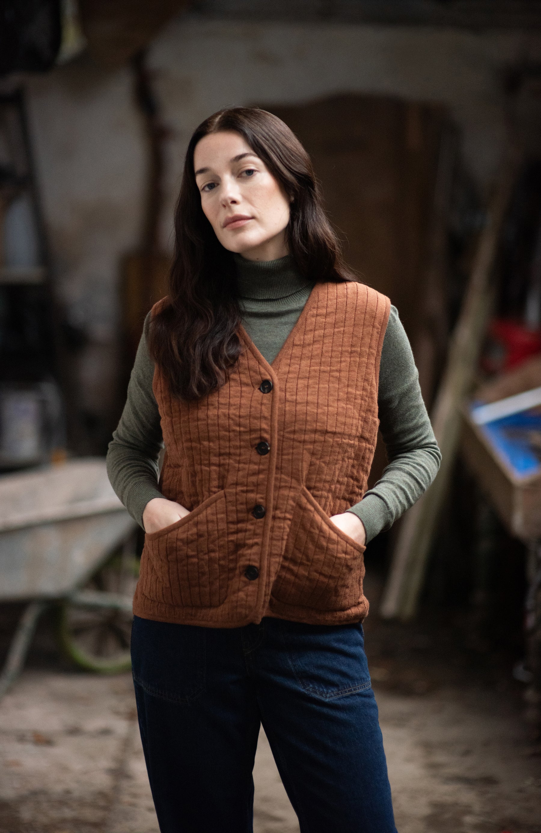 A woman wearing a rust-brown quilted button-front vest over a sage green turtleneck by Paul James Knitwear and dark blue jeans, standing in a rustic workshop environment.