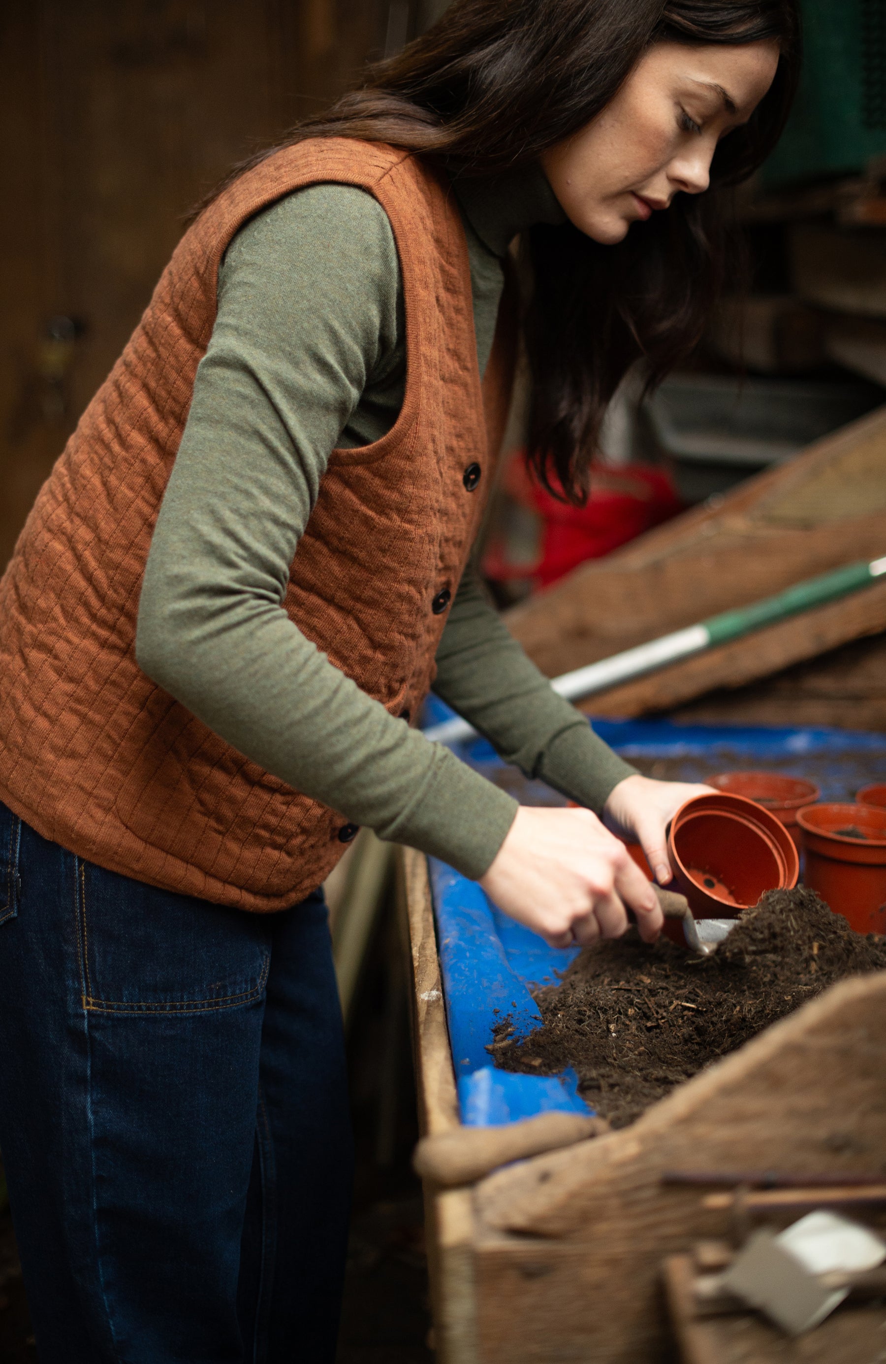 A woman wearing a brown quilted cotton vest by Paul James Knitwear over an olive green long-sleeve roll neck jumper and blue jeans is gardening, placing soil into a small terracotta pot on a blue table tray in a rustic workshop or greenhouse setting.