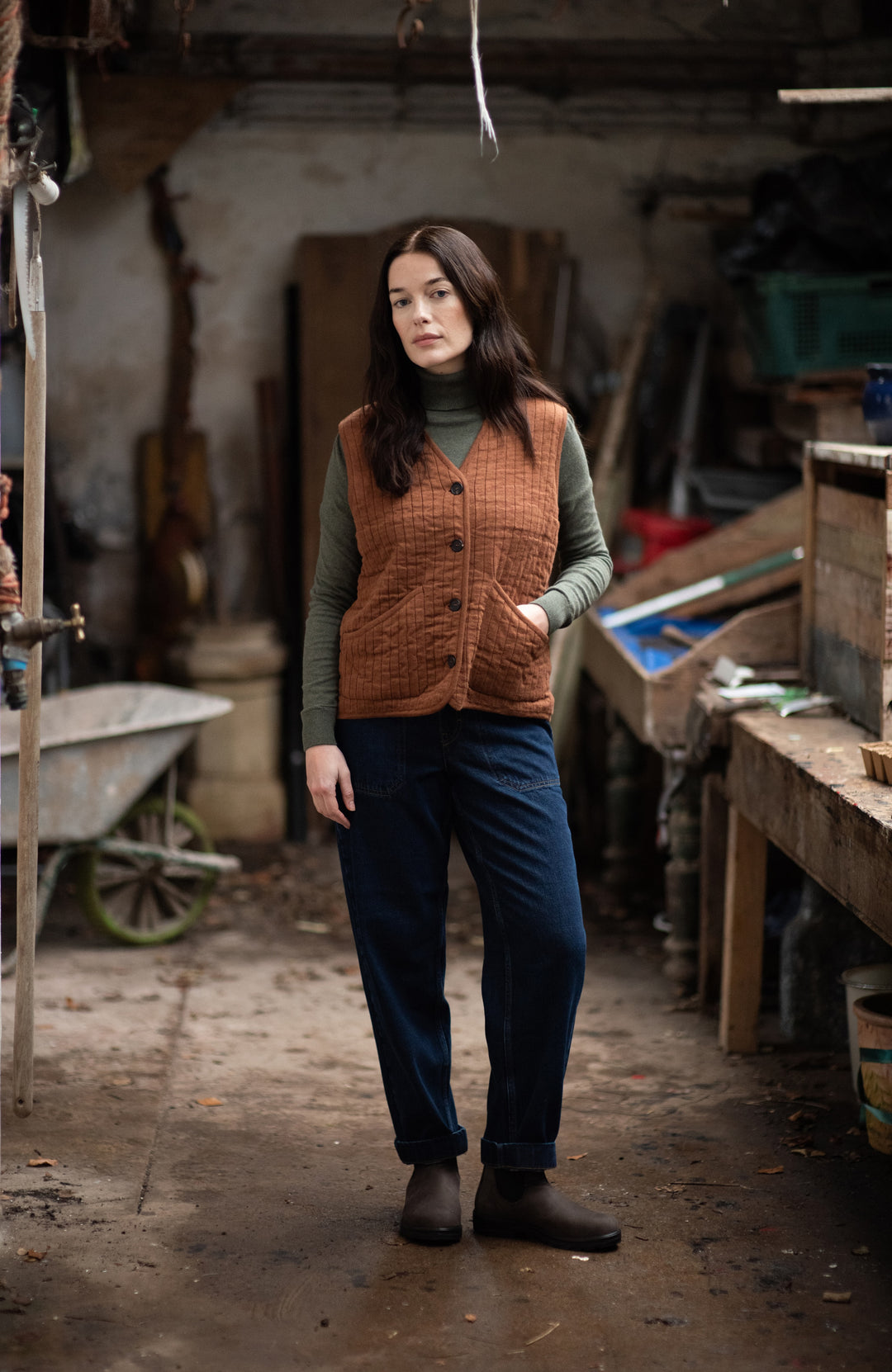 A woman stands in a rustic workshop or garage wearing a brown quilted button-front vest by paul james knitwear over an olive green turtleneck, dark blue pants, and dark boots.