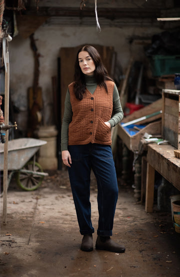 A woman stands in a rustic workshop or garage wearing a brown quilted button-front vest by paul james knitwear over an olive green turtleneck, dark blue pants, and dark boots.