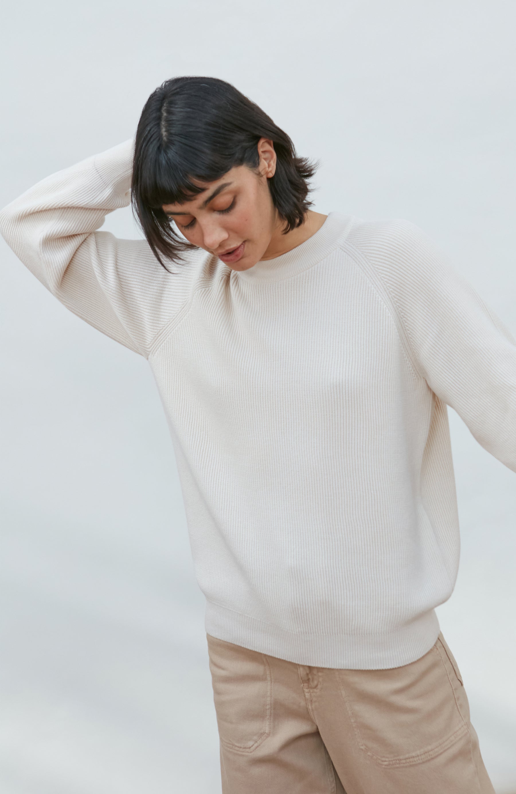 A woman wearing a cream-colored ribbed knit sweater and beige pants, posing against a light gray studio background. Made by Paul James Knitwear. 