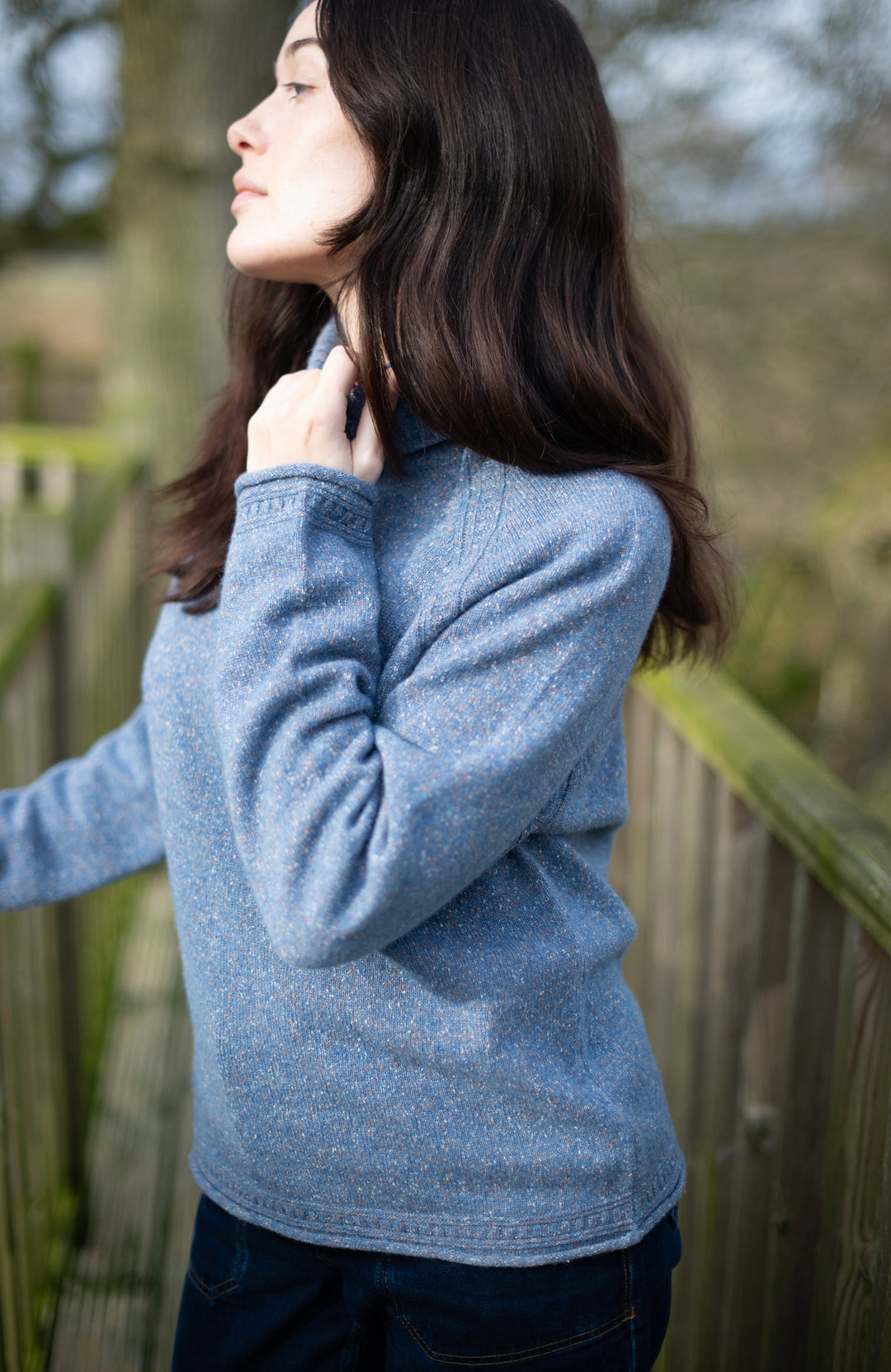A woman wearing a heathered light blue wool silk long-sleeve knit sweater by Paul James Knitwear and dark jeans, standing outdoors near a wooden fence.