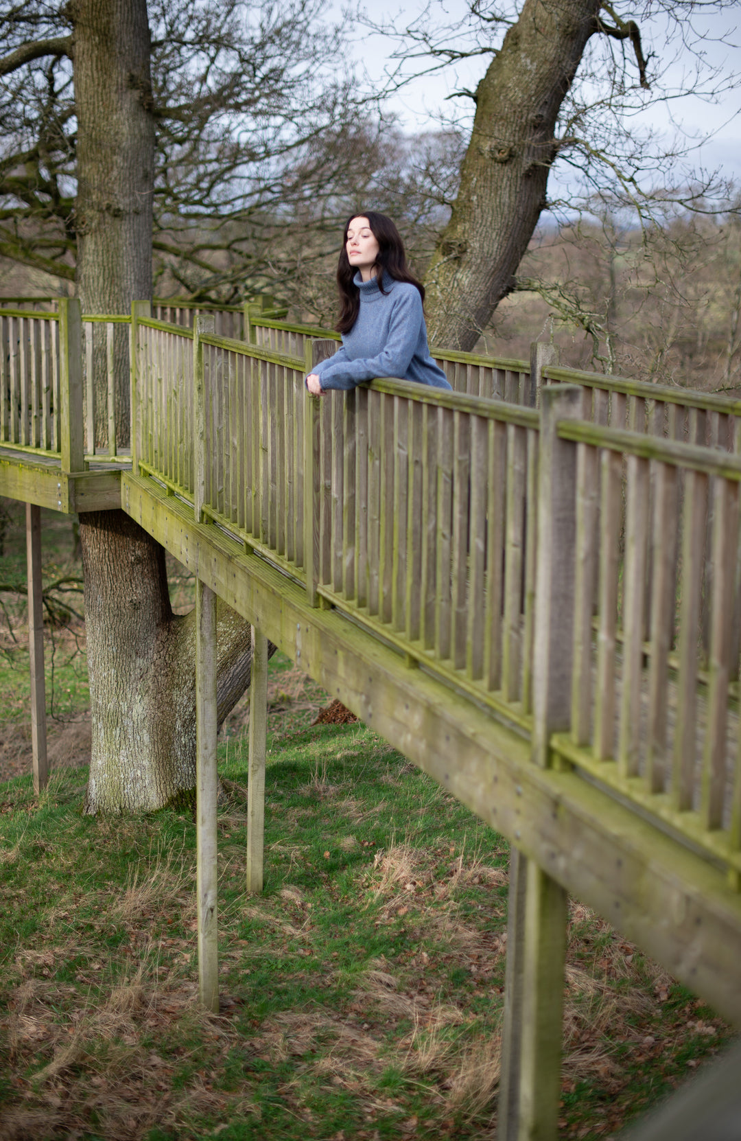 A woman wearing a heathered light blue wool silk long-sleeve knit sweater by Paul James Knitwear leans against a wooden boardwalk railing in a park with large trees and grass below.