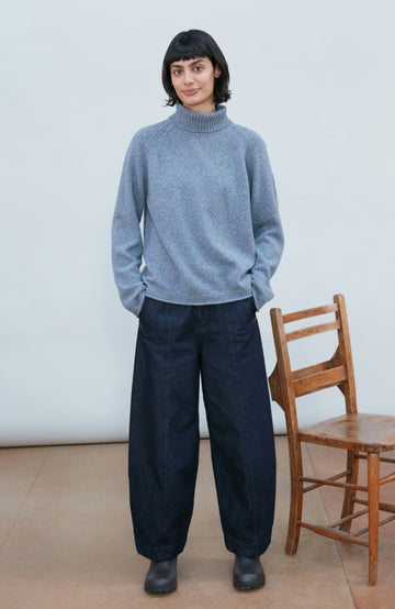 A woman with short dark hair wearing a textured wool silk heathered blue-gray turtleneck sweater Paul James Knitwear, photographed against a clean light gray background.
