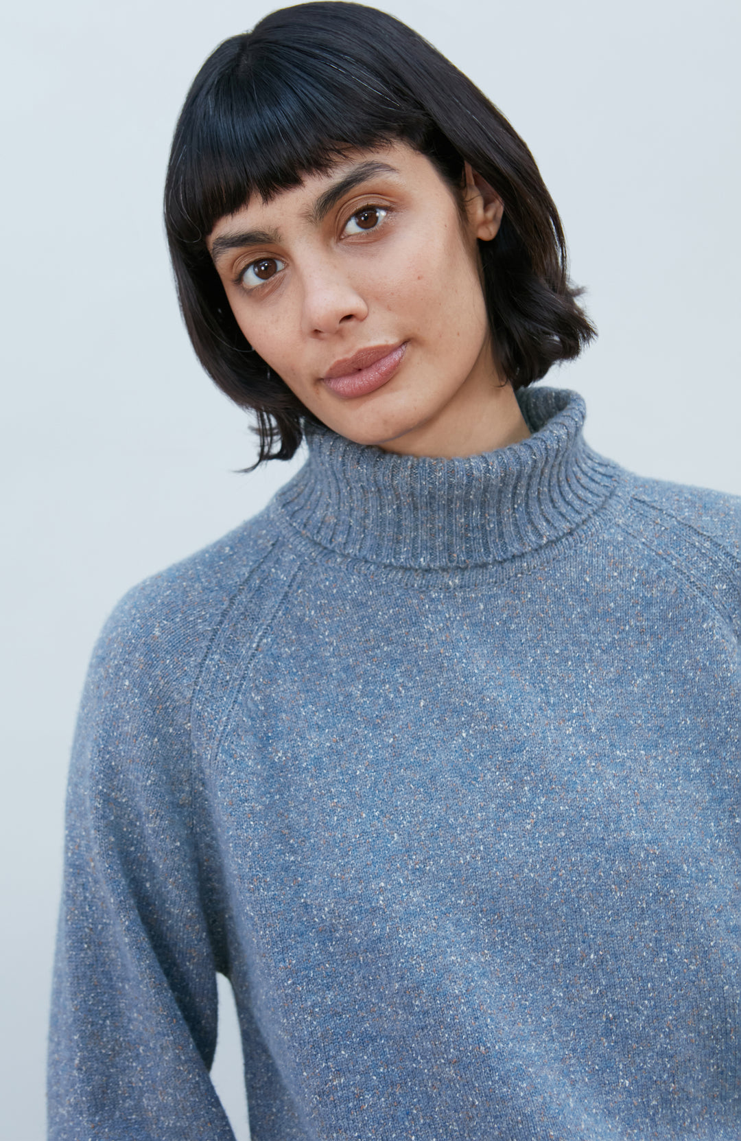 A woman with short dark hair and bangs wearing a textured wool silk heathered blue-gray turtleneck sweater Paul James Knitwear, photographed against a clean light gray background.