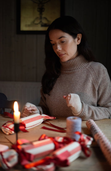 A woman in a beige wool turtleneck sweater by Paul James Knitwear sits at a wooden table, wrapping a red and white striped paper ornament with red ribbon. A lit candle and other wrapped ornaments are on the table in a dimly lit room.