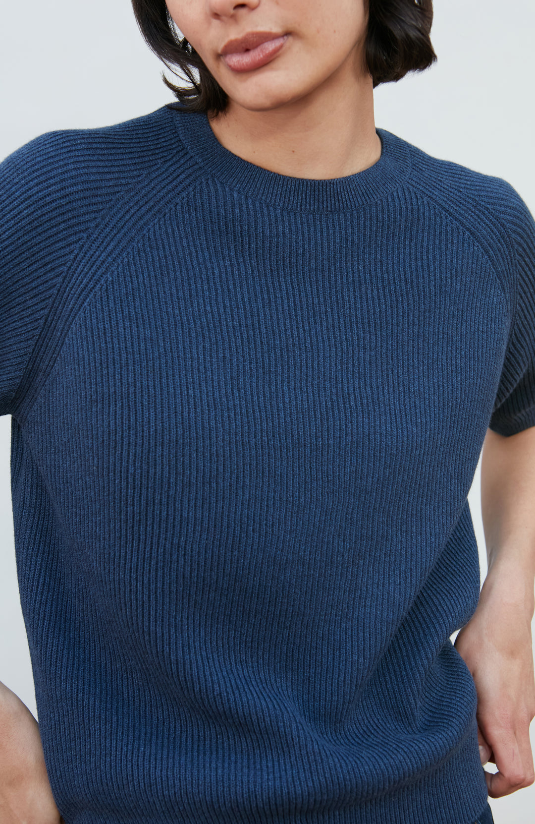 A woman wearing a dark navy blue ribbed short-sleeve mock neck top by paul james knitwear and dark blue jeans, posing against a light gray background.