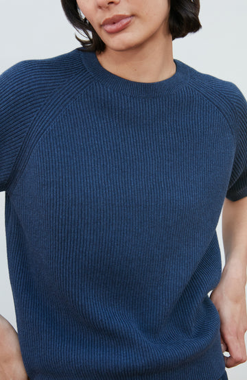 A woman wearing a dark navy blue ribbed short-sleeve mock neck top by paul james knitwear and dark blue jeans, posing against a light gray background.