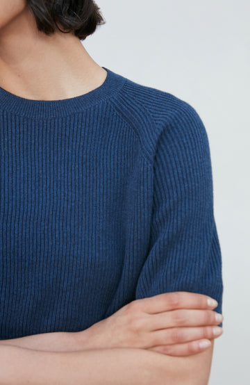 A woman wearing a dark navy blue ribbed short-sleeve mock neck top by paul james knitwear and dark blue jeans, posing against a light gray background.