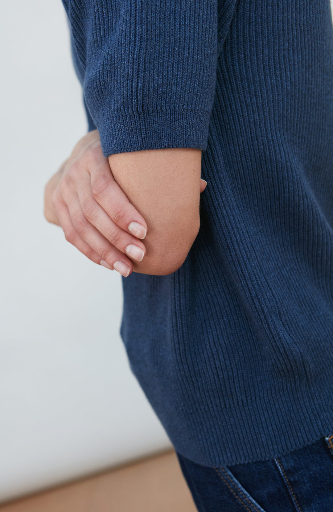 A woman wearing a dark navy blue ribbed short-sleeve mock neck top by paul james knitwear and dark blue jeans, posing against a light gray background.