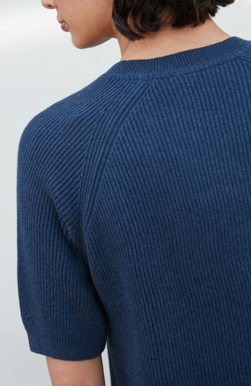 A woman wearing a dark navy blue ribbed short-sleeve mock neck top by paul james knitwear and dark blue jeans, posing against a light gray background.