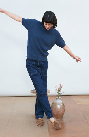 A woman wearing a dark navy blue ribbed short-sleeve mock neck top by paul james knitwear and dark blue jeans, posing against a light gray background.