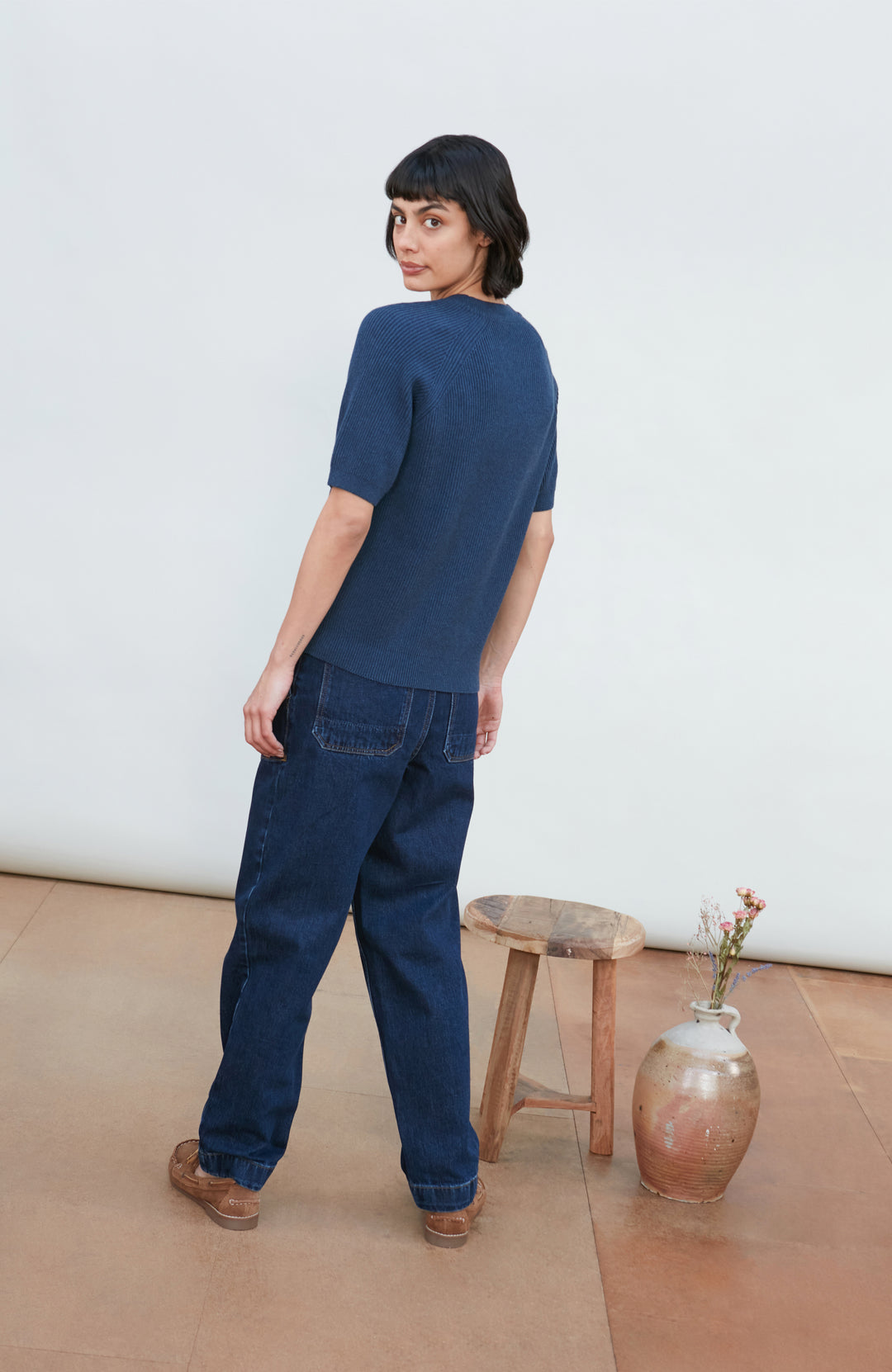 A woman wearing a dark navy blue ribbed short-sleeve mock neck top by paul james knitwear and dark blue jeans, posing against a light gray background.