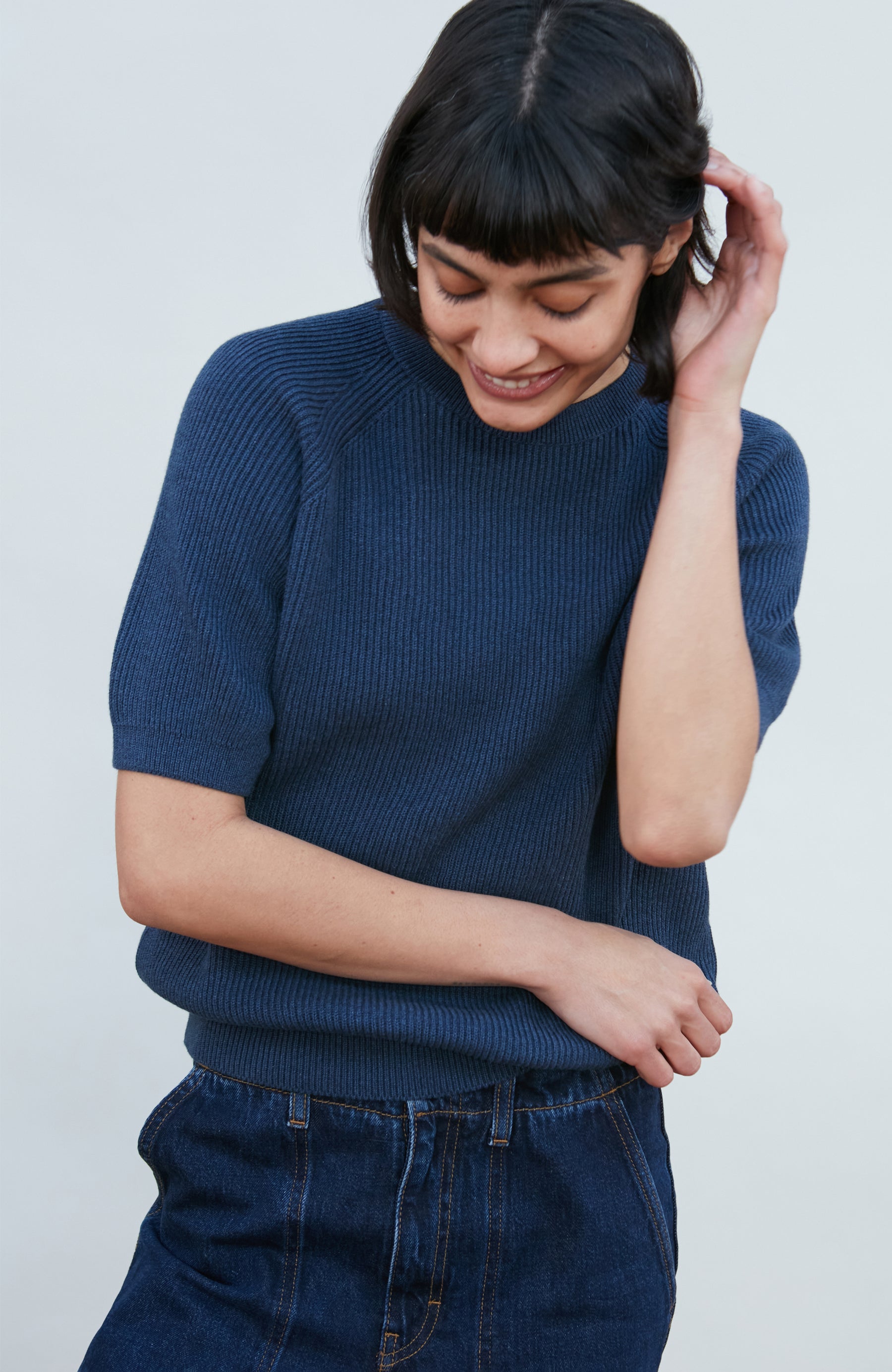 A woman wearing a dark navy blue ribbed short-sleeve mock neck top by paul james knitwear and dark blue jeans, posing against a light gray background.