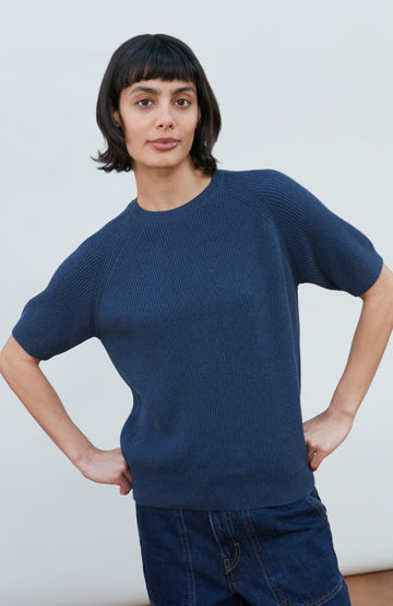 A woman wearing a dark navy blue ribbed short-sleeve mock neck top by paul james knitwear and dark blue jeans, posing against a light gray background.