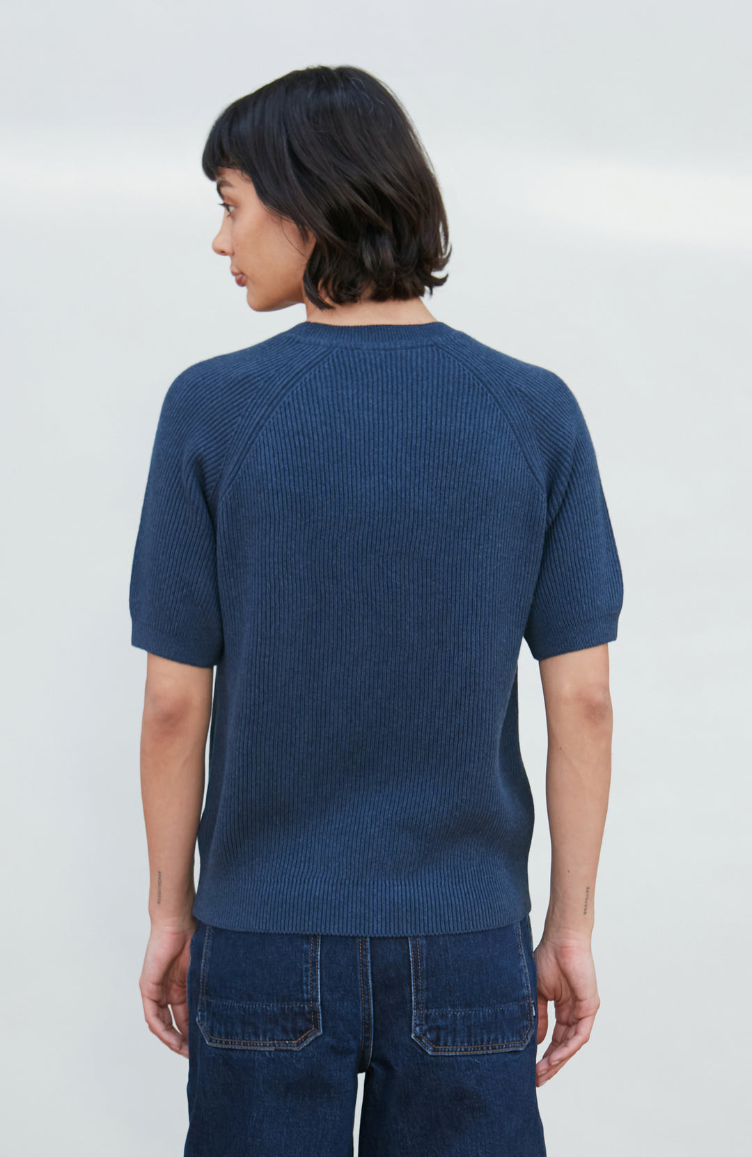 A woman wearing a dark navy blue ribbed short-sleeve mock neck top by paul james knitwear and dark blue jeans, posing against a light gray background.
